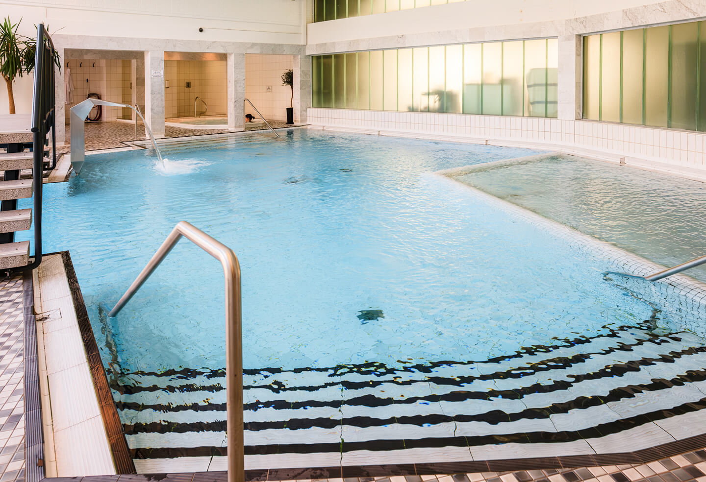 Großer Innenpool mit Wasserschwall auf der linken Seite, kleinerem Becken rechts und breiter Treppe mit Geländer zum Hineingehen im Vordergund. Im Hinergrund befindet sich ein separater Bereich mit Whirlpool.