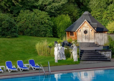 Garten mit Poolbereich und Außensauna. Vor der kleinen Hütte mit Sauna stehen drei Skulpturen, am Pool stehen blaue Poolliegen.
