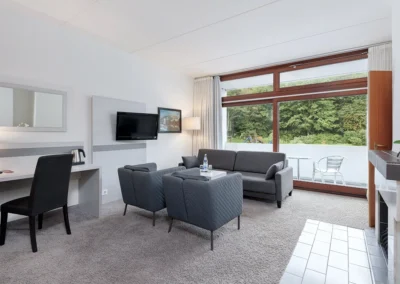 Hotelzimmer mit einem Sofa, zwei Sesseln und Fernseher an der Wand vor großer Fensterfront mit Blick auf einen Balkon. Rechts befindet sich ein Kamin, gegenüber steht ein Schreibtisch mit Polsterstuhl.