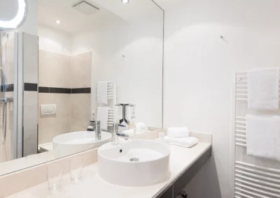 Badezimmer mit Waschtisch vor großem Wandspiegel. Rechts daneben an der Wand ein Heizkörper mit Handtüchern.