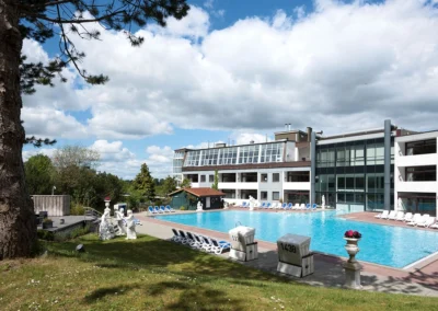 Blick aus dem Garten auf den Pool und das Hotel. Um den Pool herum stehen Poolliegen und Strandkörbe.
