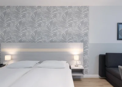 Doppelbett mit Nachttischen und Nachttischlampen vor einer Wand mit grau-weißer Deko-Tapete mit Palmenblättern. Rechts die Ecke eines dunkelgrauen Sofas neben dem Bett.