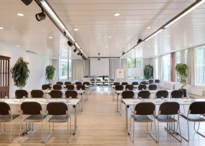 Tagungsraum mit mehreren Tisch- und Stuhlreihen. Ausgerichtet auf einen Tisch mit Stühlen vor einer von der Decke hängenden Leinwand.
