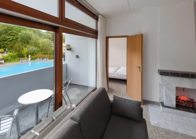 Blick über ein graues Sofa durch die offene Tür ins Schlafzimmer. Rechts ein Kamin und links großer Balkon mit Balkonmöbeln und Aussicht auf den Pool.