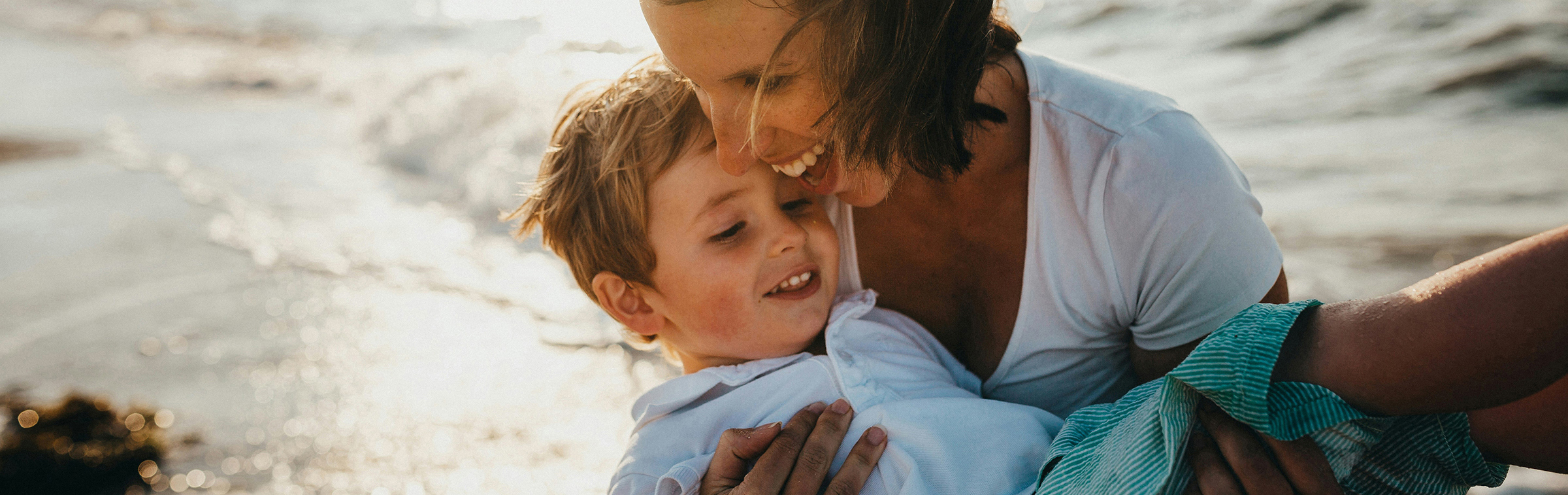 Eine lachende Frau trägt einen Jungen an einem Strand auf den Armen.