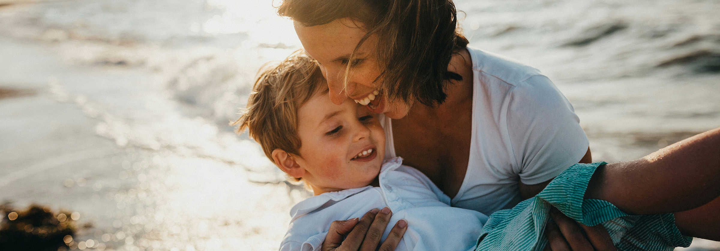 Eine lachende Frau trägt einen Jungen an einem Strand auf den Armen.